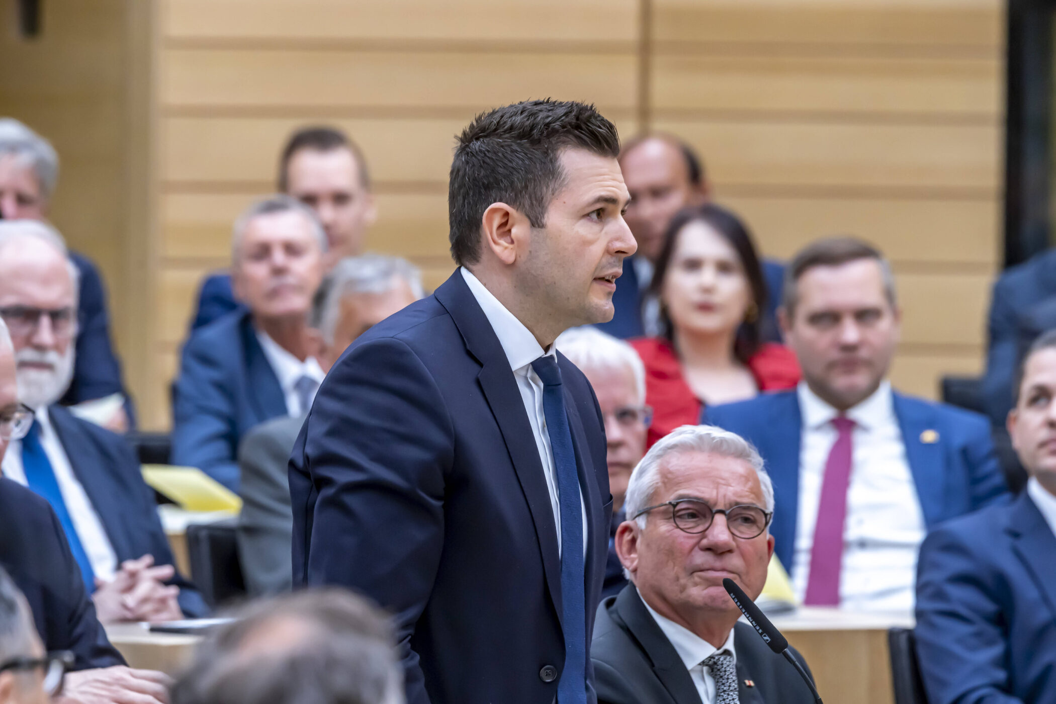 Mann in Anzug spricht im Raum, umgeben von anderen Männern im Anzug.