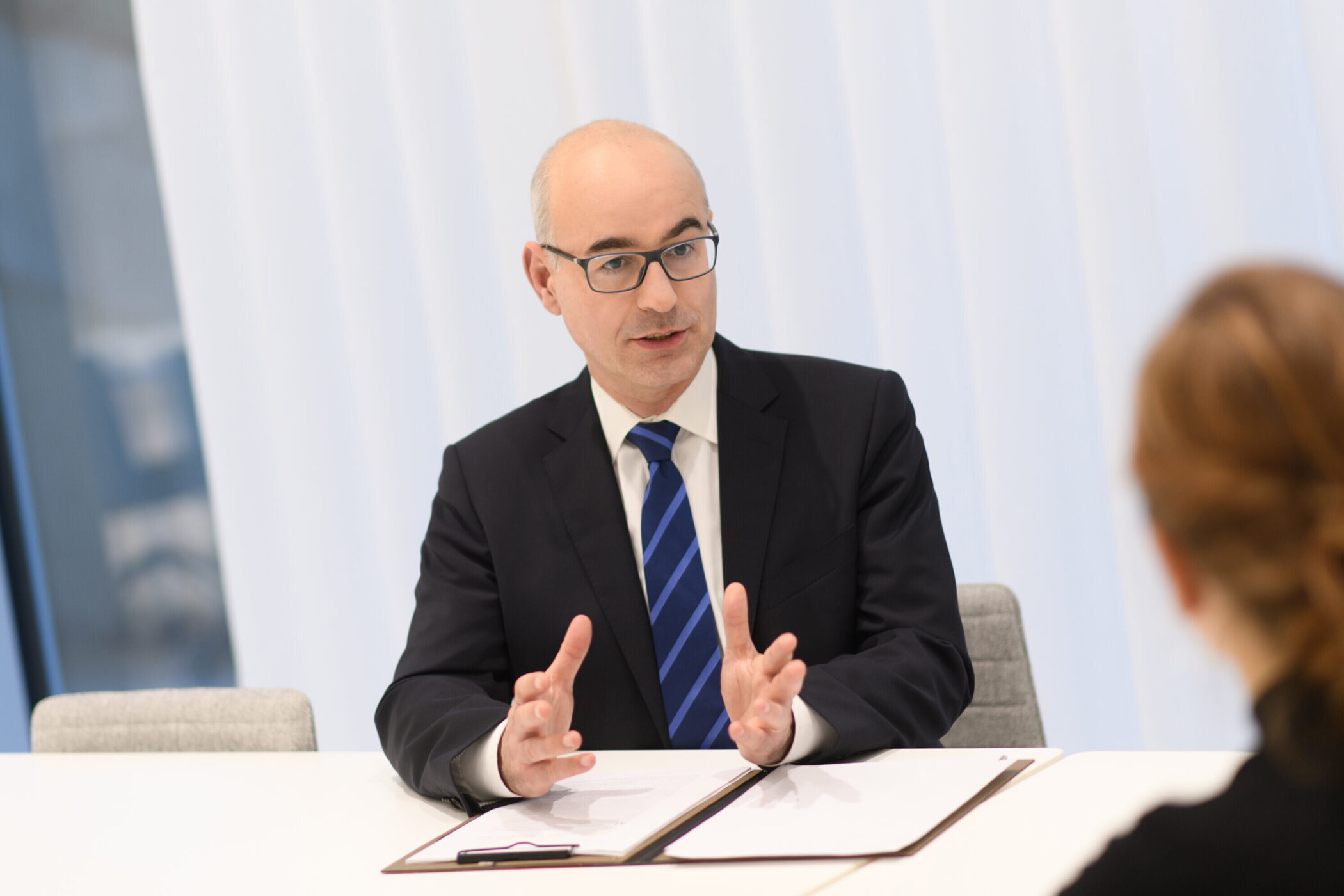 Zwei Personen an einem Tisch, eine hält einen Stift, helle Umgebung.