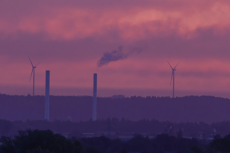Zwei Windräder und zwei Schornsteine vor einem rosa Himmel.
