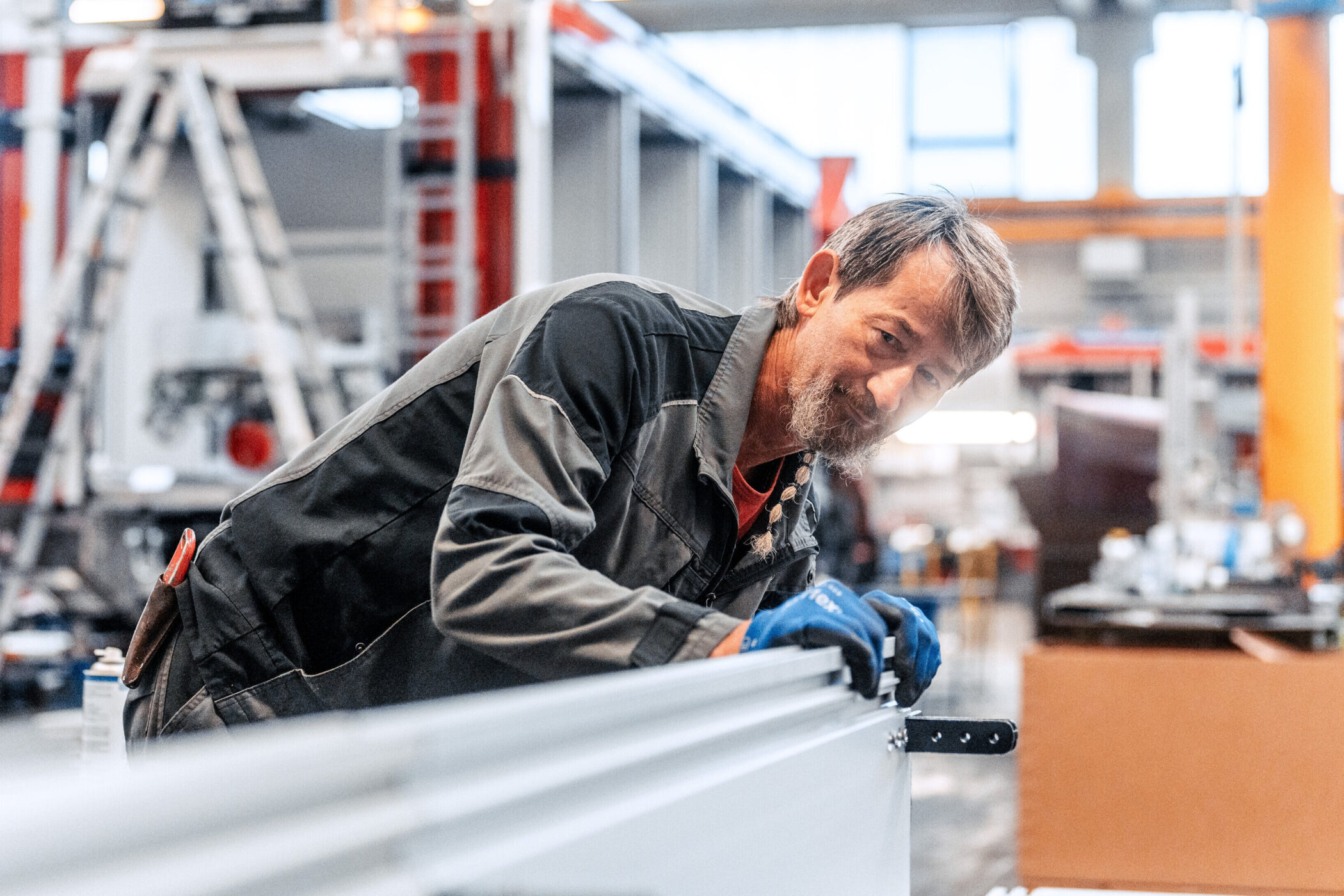 Person in Arbeitskleidung lehnt sich über Metallstruktur in Werkshalle.