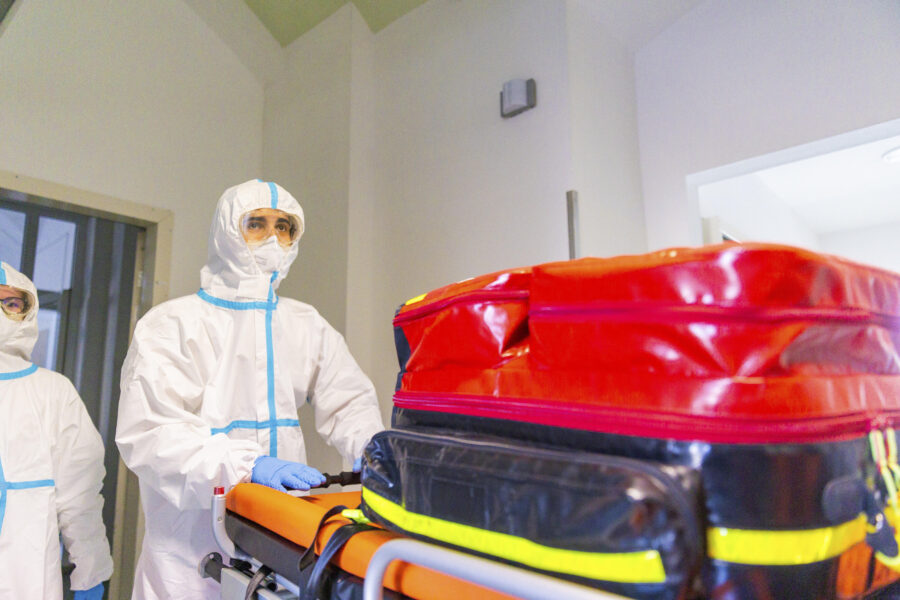 Zwei Personen in Schutzanzügen bewegen eine Trage mit roter Tasche.