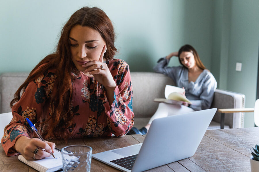 Zwei Frauen, eine schreibt, eine liest, sitzen in einem Raum mit Laptop auf dem Tisch.