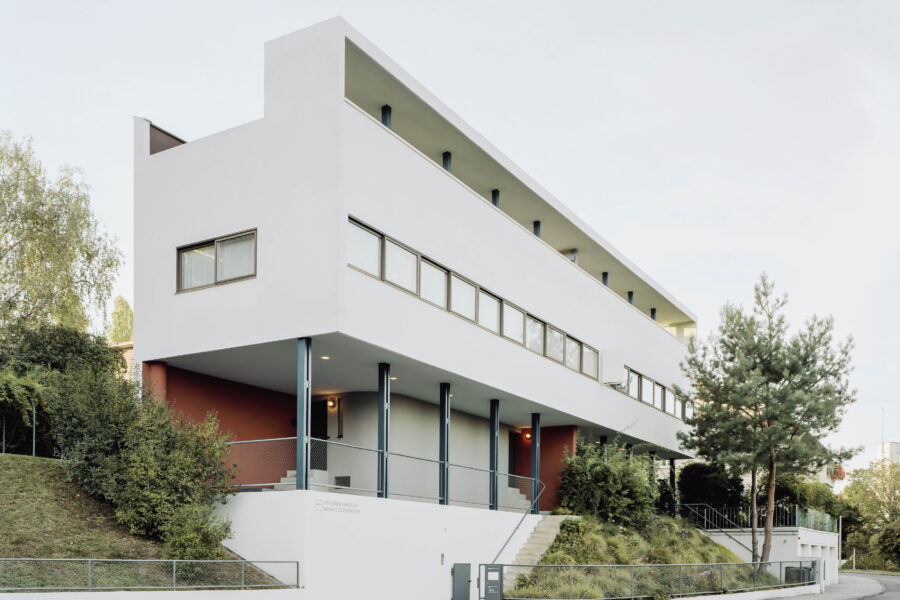Modernes, weißes Gebäude mit vielen Fenstern, Treppe führt zum Eingang, Bäume daneben.