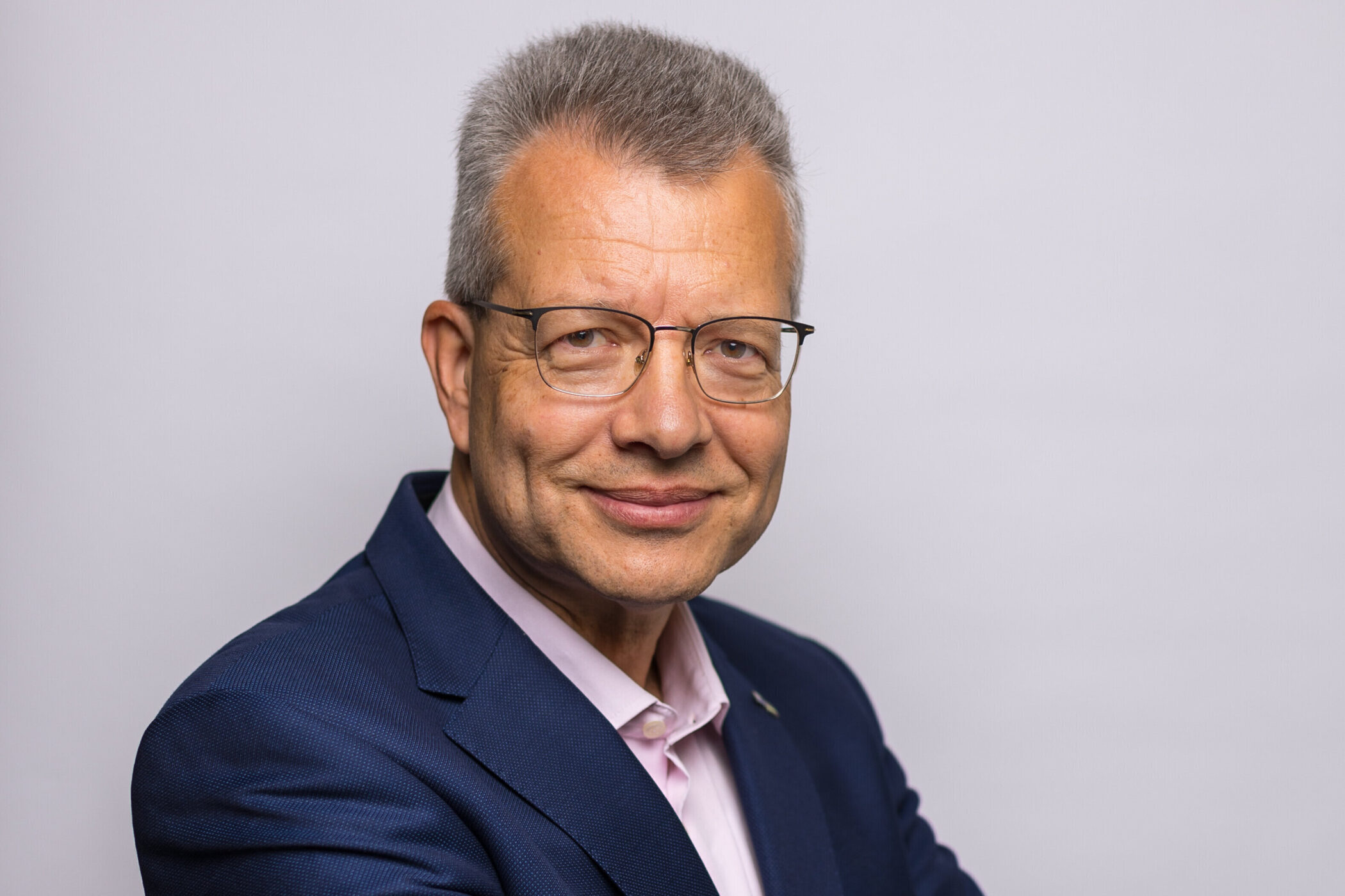 Älterer Mann mit Brille im Anzug lächelt vor einem hellen Hintergrund.