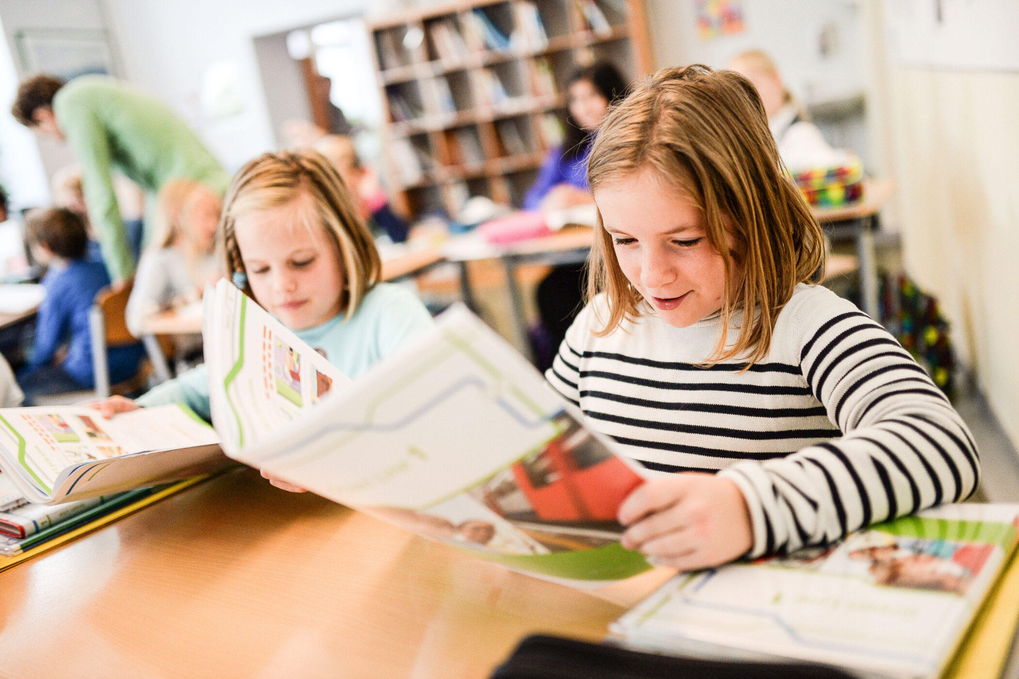 Zwei Kinder lesen in einem Klassenzimmer Bücher. Im Hintergrund Schüler an Tischen.