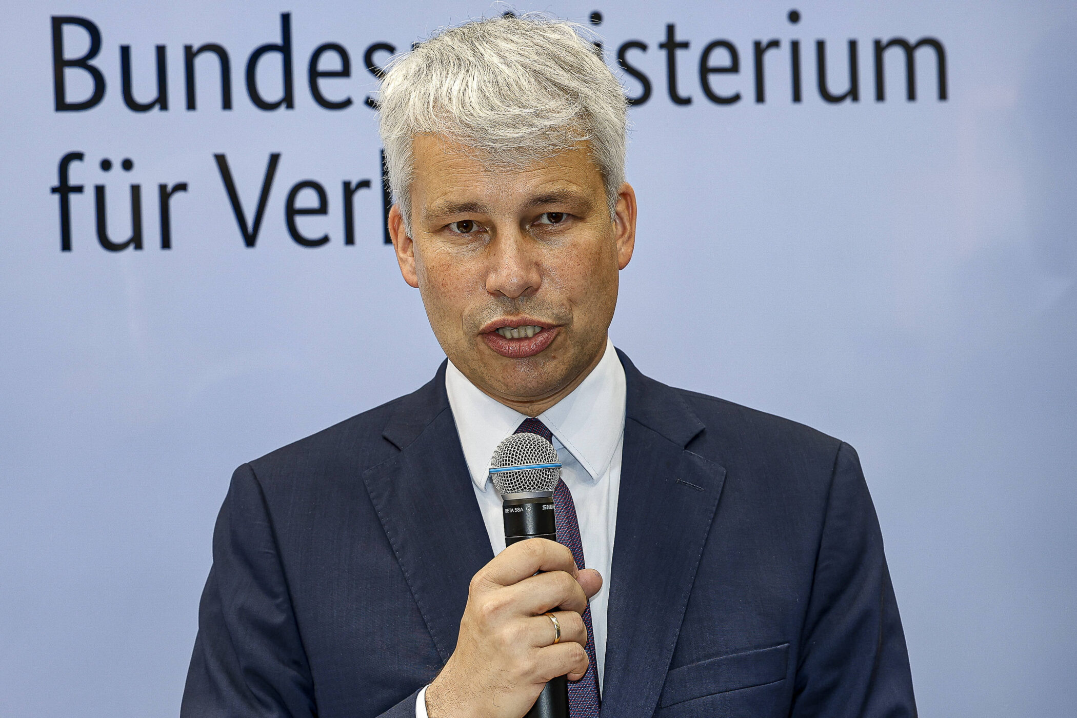 Mann im Anzug mit Mikrofon vor Schriftzug "Bundesministerium für Ver".