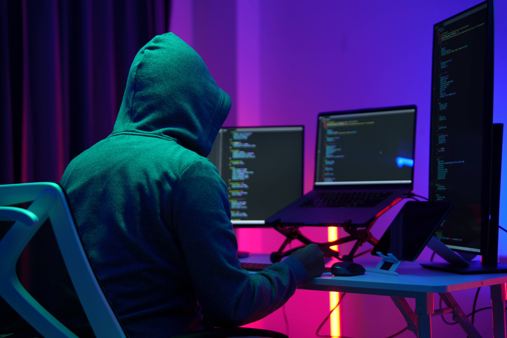 Person mit Kapuzenpulli sitzt vor mehreren Monitoren mit Code.