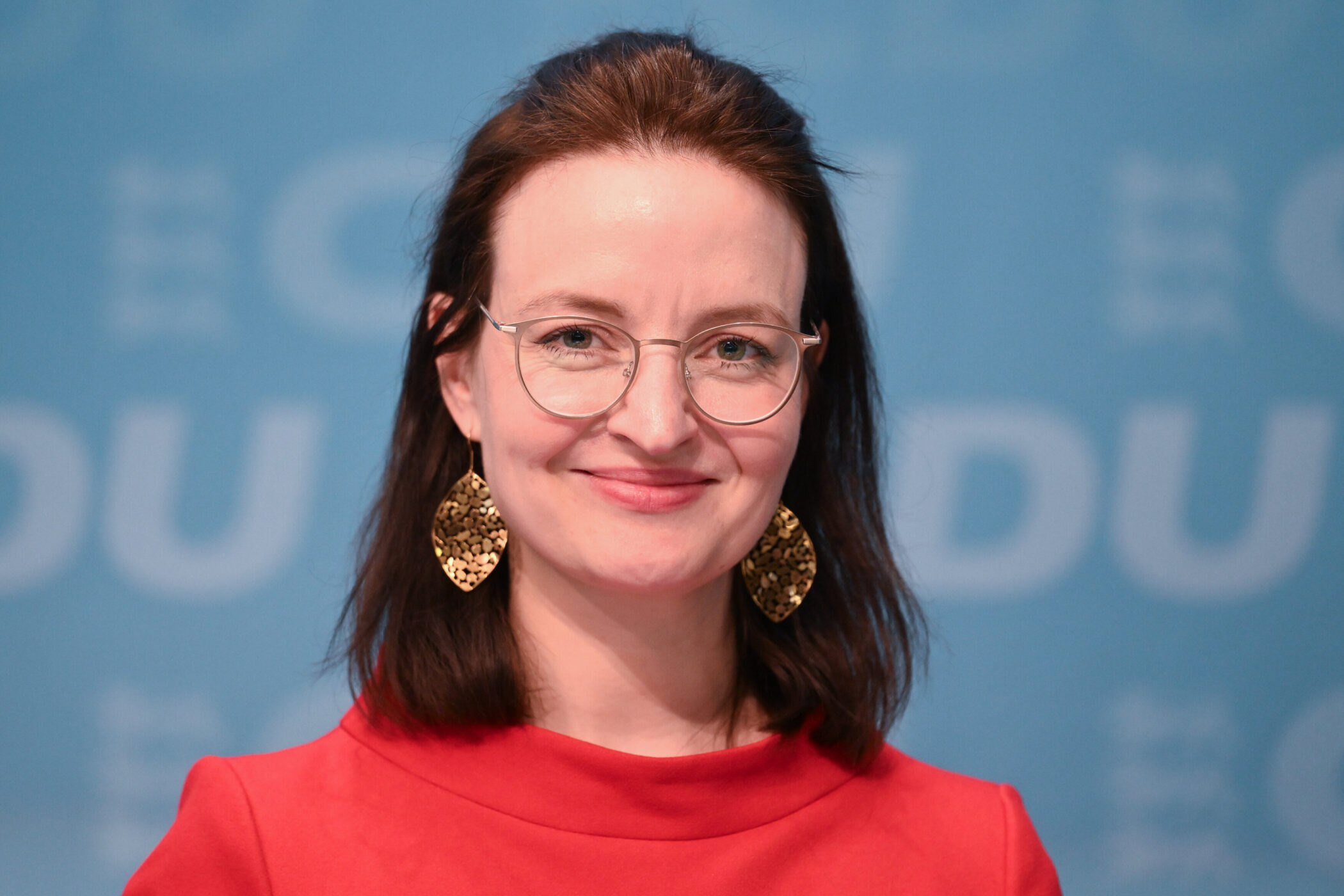 Eine Person in rotem Oberteil mit Brille und goldenen Ohrringen vor blauem Hintergrund.