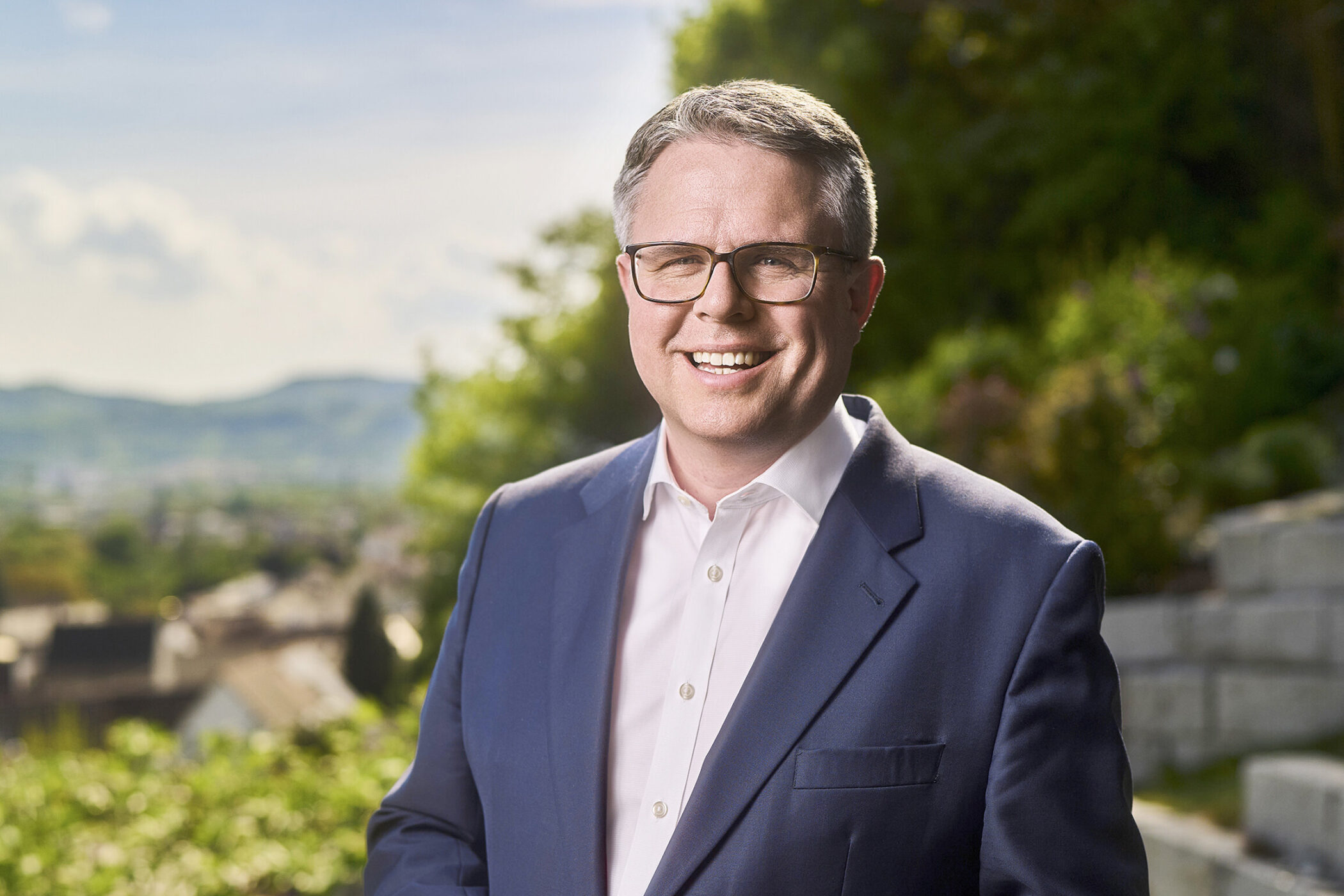 Mann in Anzug, lächelt, Brille tragend, draußen vor unscharfer Landschaft.