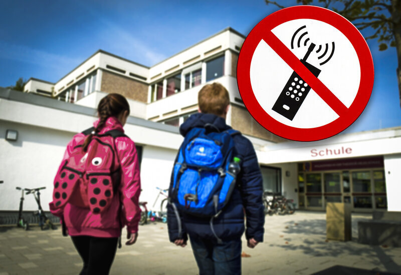 Zwei Kinder mit Rucksäcken gehen zur Schule. Handy-Verbotsschild über dem Eingang.