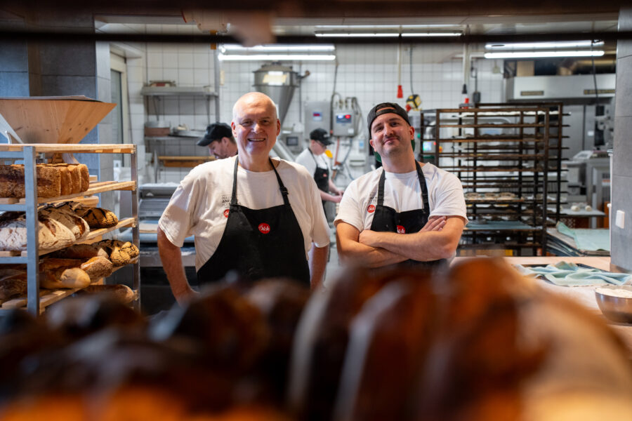 Zwei Menschen in einer Bäckerei, Brote auf einem Regal im Hintergrund.