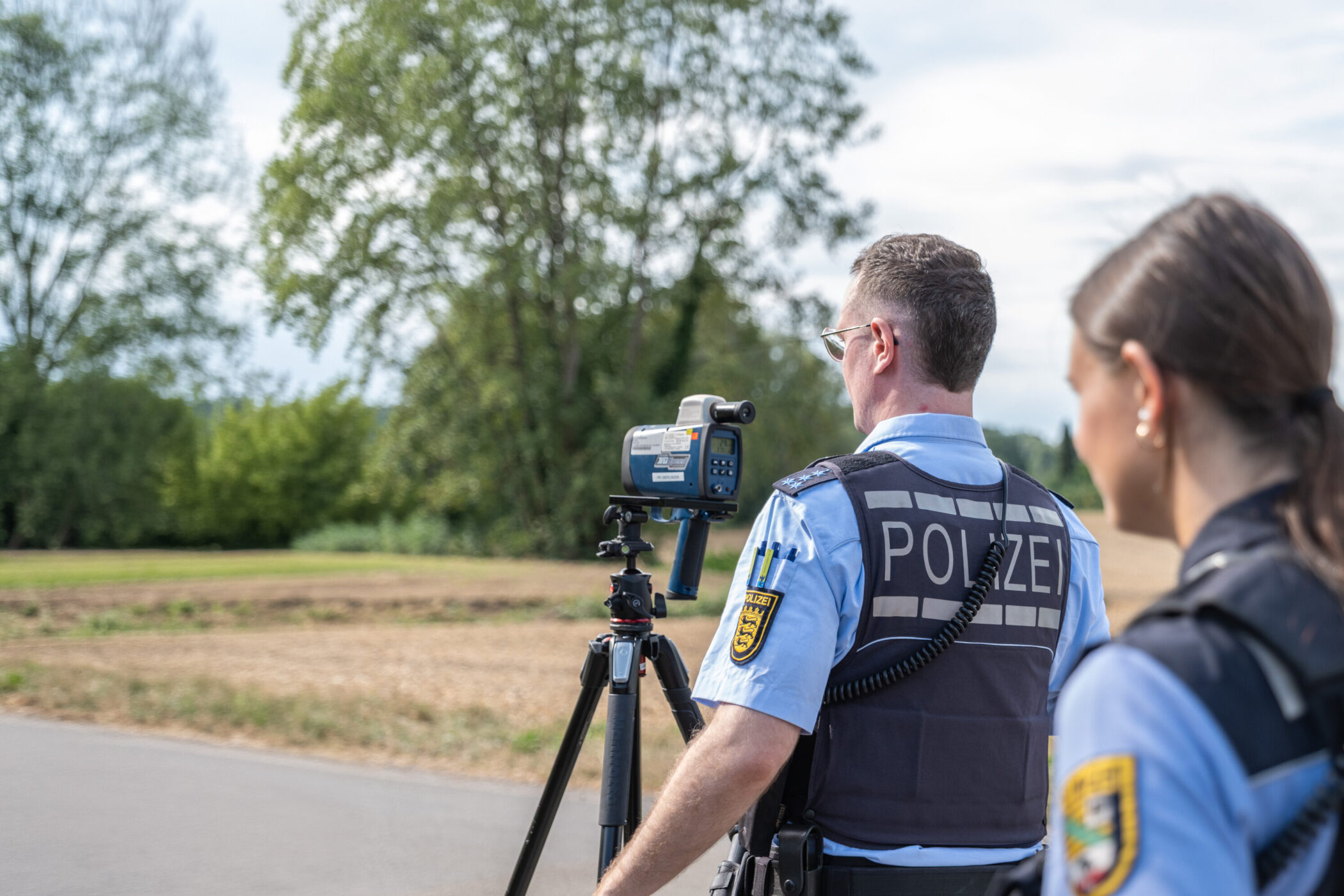 Zwei Polizisten überwachen mit einem Gerät eine Straße, im Hintergrund Bäume.
