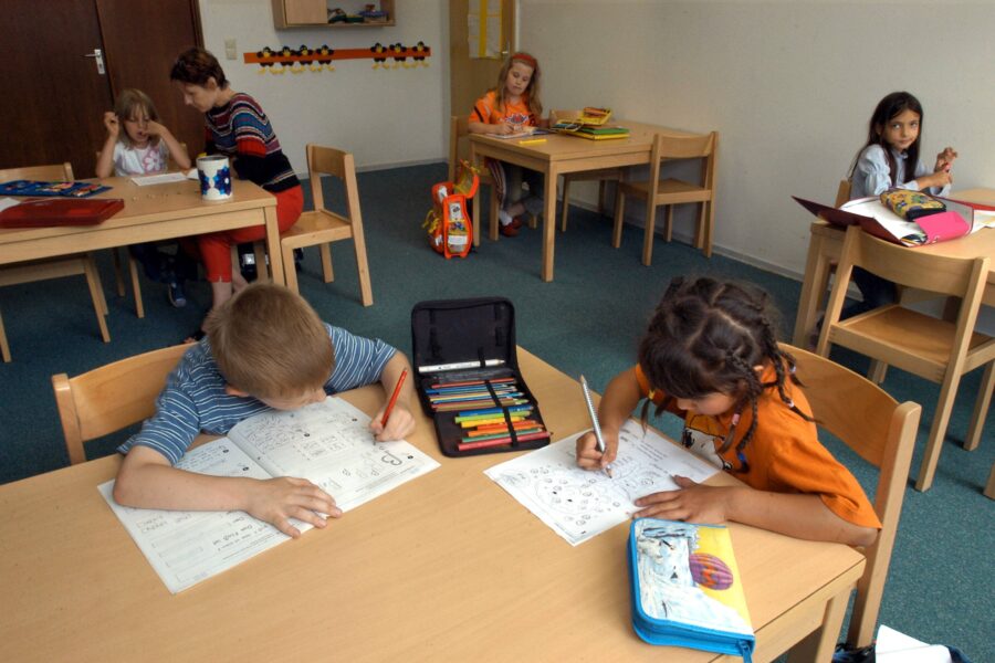 Mehrere Kinder an Tischen beim Schreiben, Lehrerin hilft einem Kind.