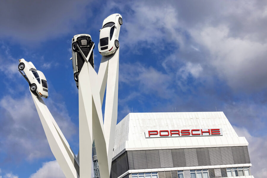 Drei weiße Autos auf hohen Stützen, Gebäude im Hintergrund mit Schrift "Porsche".