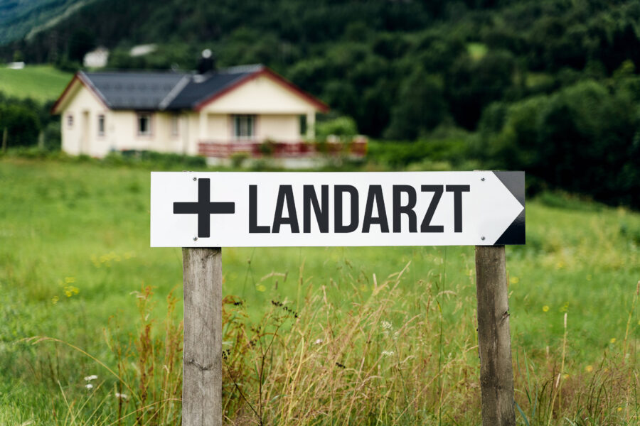 Schild mit Aufschrift "Landarzt" und Pfeil in ländlicher Umgebung.