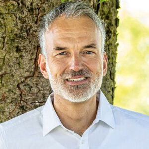 Ein Mann mit weißem Hemd steht vor einem Baum.