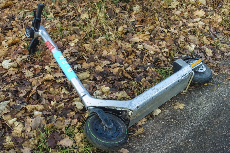 Silberner E-Scooter liegt auf dem Rücken zwischen Herbstlaub und Gehweg.