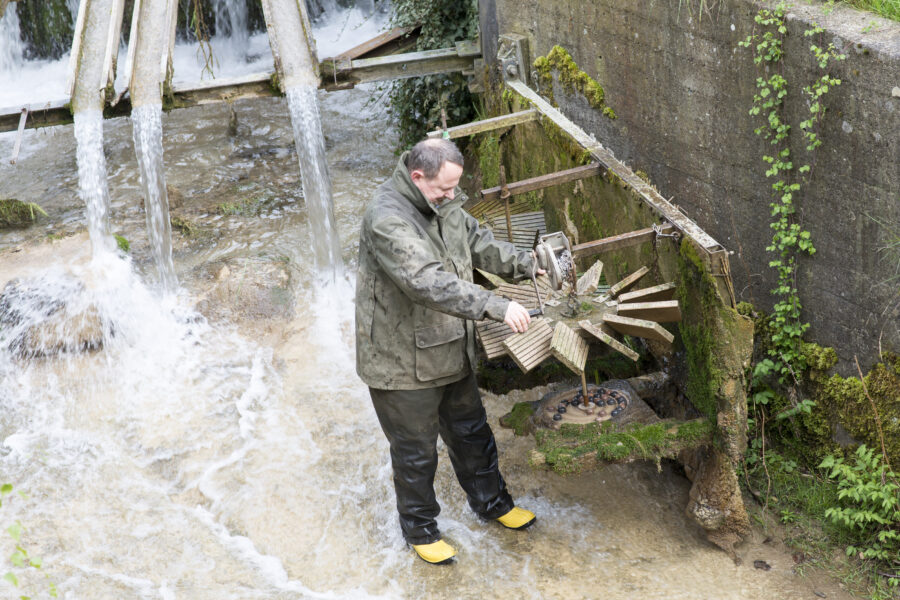 Person in Regenkleidung arbeitet an einem hölzernen Wasserrad, umgeben von fließendem Wasser.