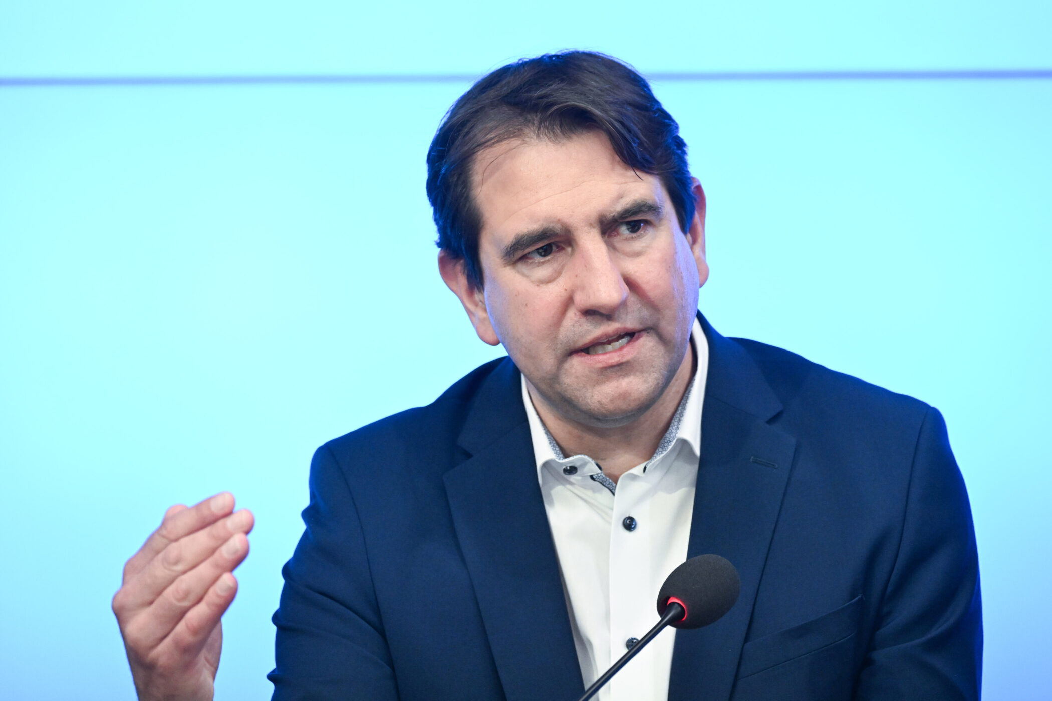 Mann spricht in Mikrofon, trägt dunklen Anzug, blauen Hintergrund.