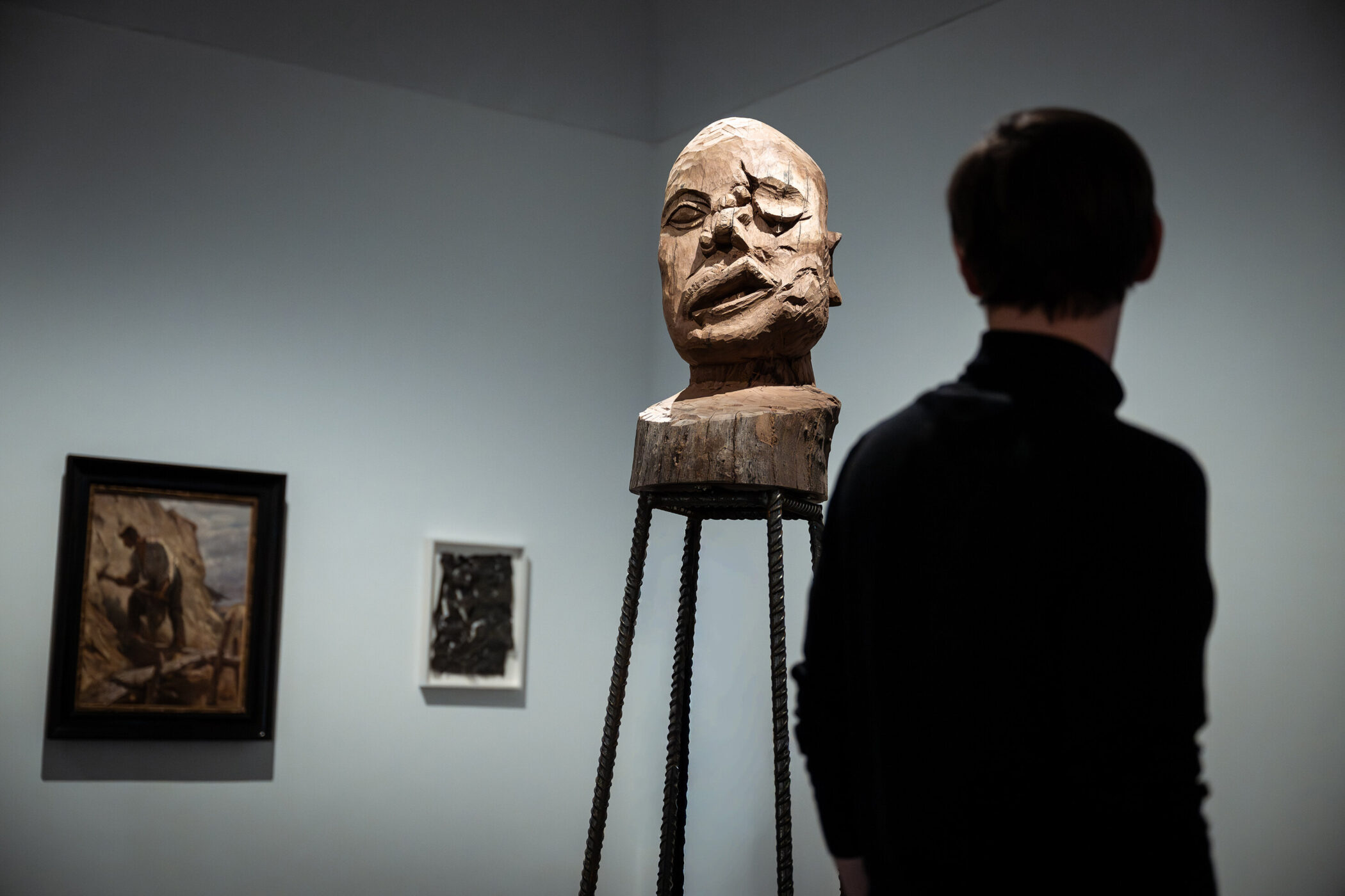Person steht in Galerie vor Holzskulptur, zwei Gemälde an der Wand.