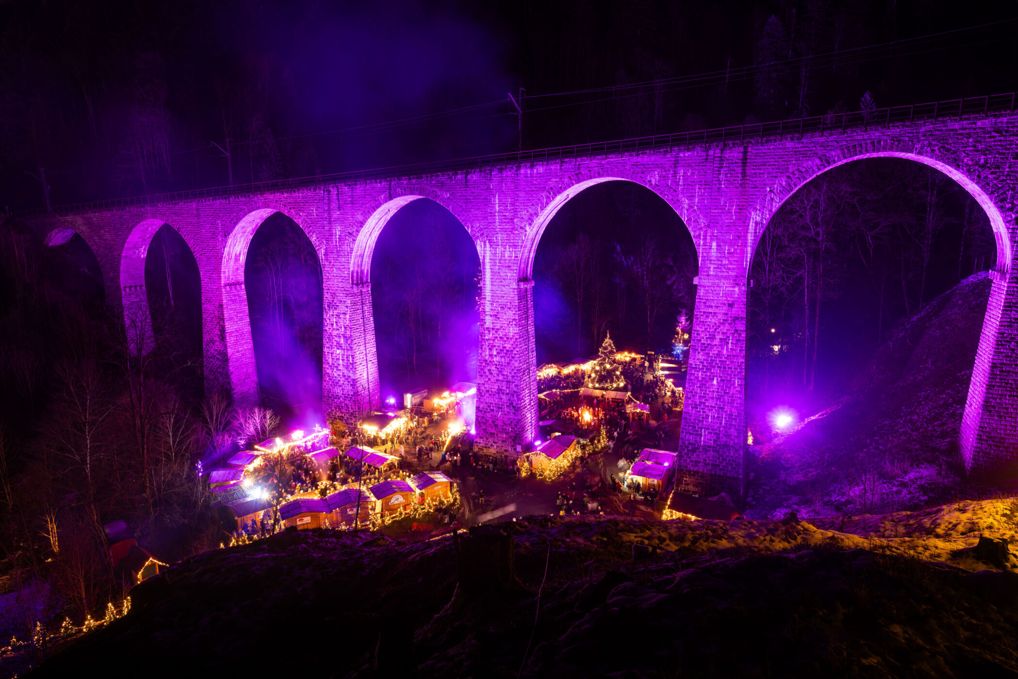 Viadukt bei Nacht in lila beleuchtet, Marktstände darunter, dunkler Wald im Hintergrund.