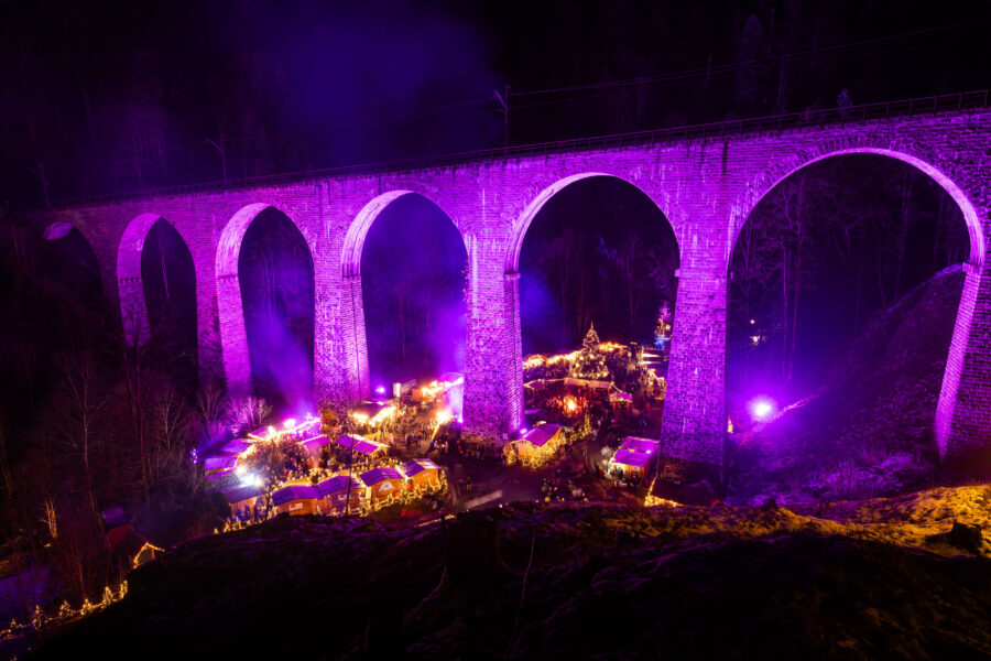 Viadukt bei Nacht in lila beleuchtet, Marktstände darunter, dunkler Wald im Hintergrund.