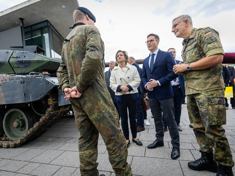 Eine Gruppe von Menschen steht vor einem Panzer, zwei in Militäruniform.