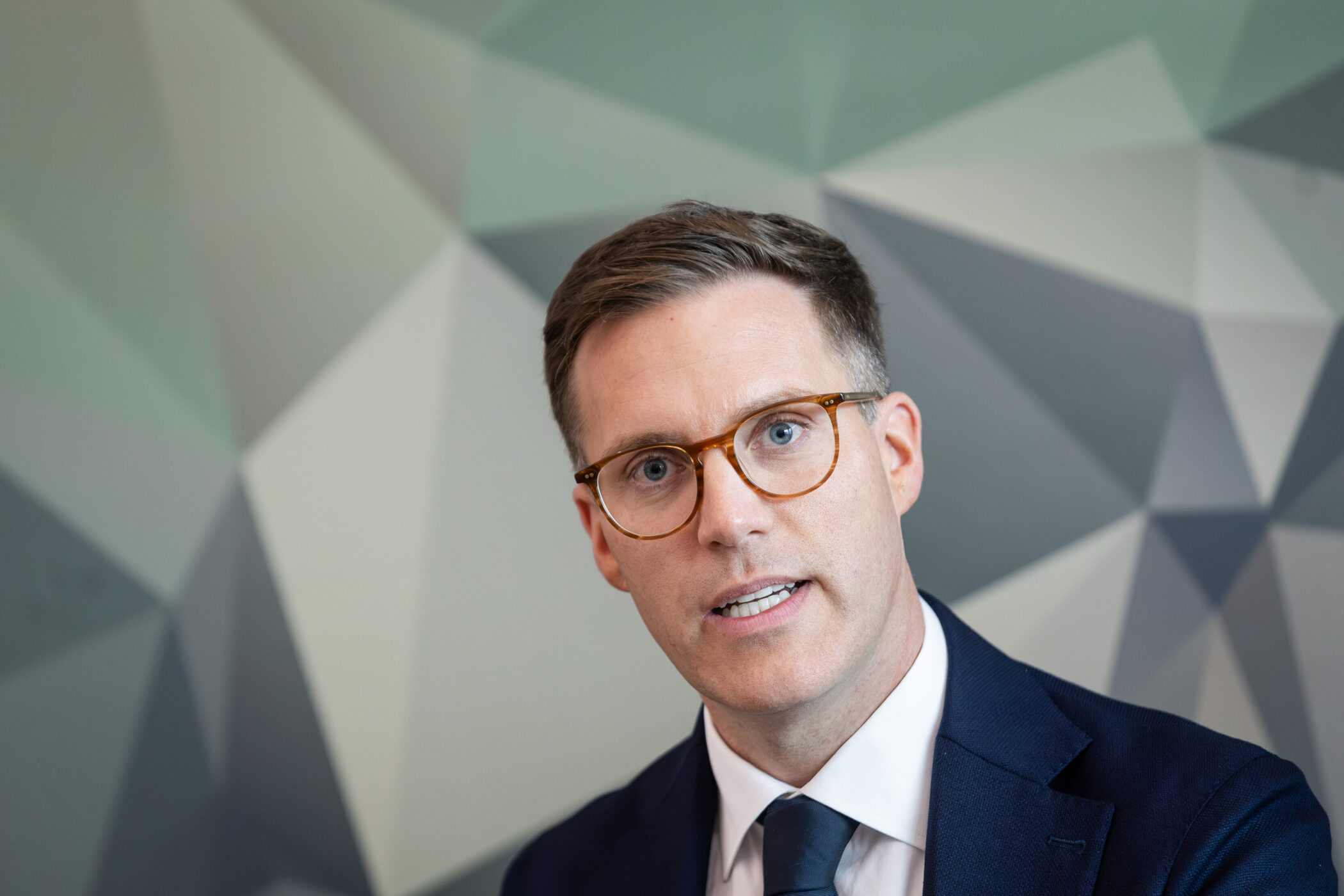 Ein Mann mit Brille und Anzug vor geometrischem Hintergrund.