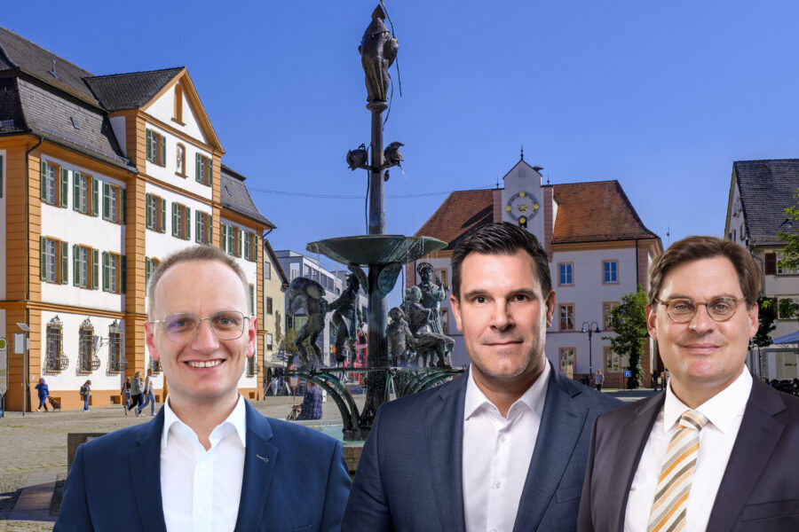 Drei Männer im Anzug vor historischem Brunnen und Gebäuden bei klarem Himmel.