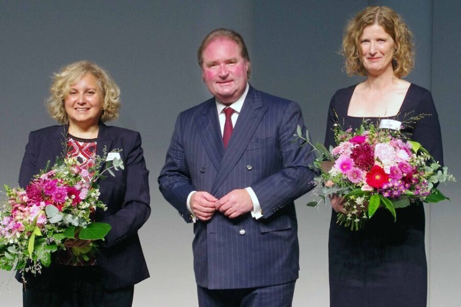 Zwei Frauen mit Blumensträußen neben einem Mann im Anzug, alle lächeln.