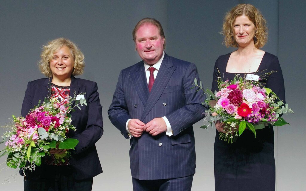 Zwei Frauen mit Blumensträußen neben einem Mann im Anzug, alle lächeln.