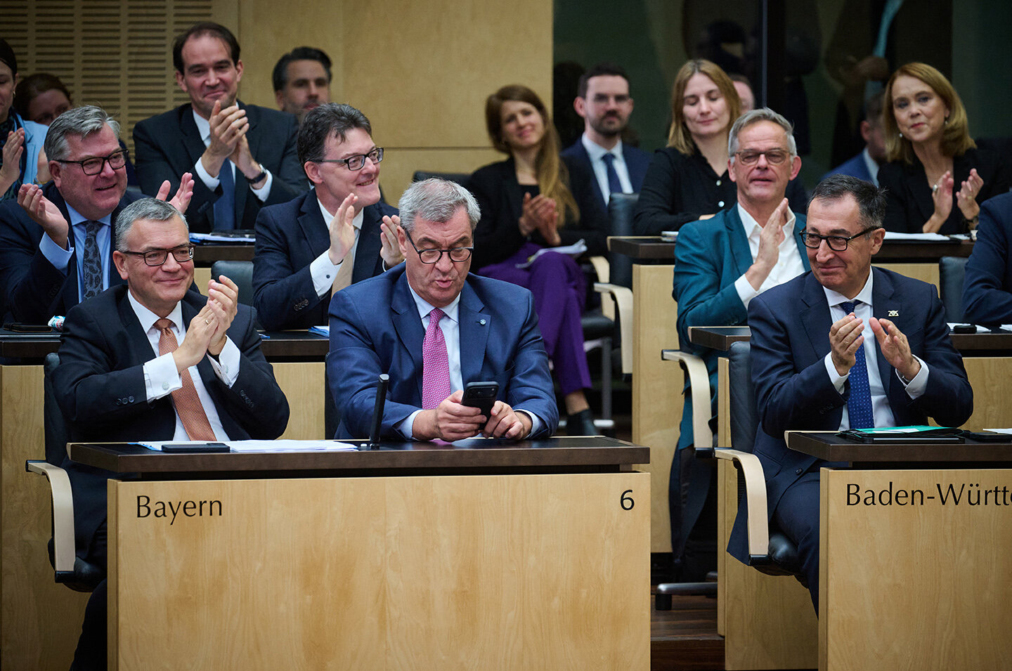 Mehrere Personen in Anzügen klatschen in einem Saal, vorne ein Mann mit Mobiltelefon.