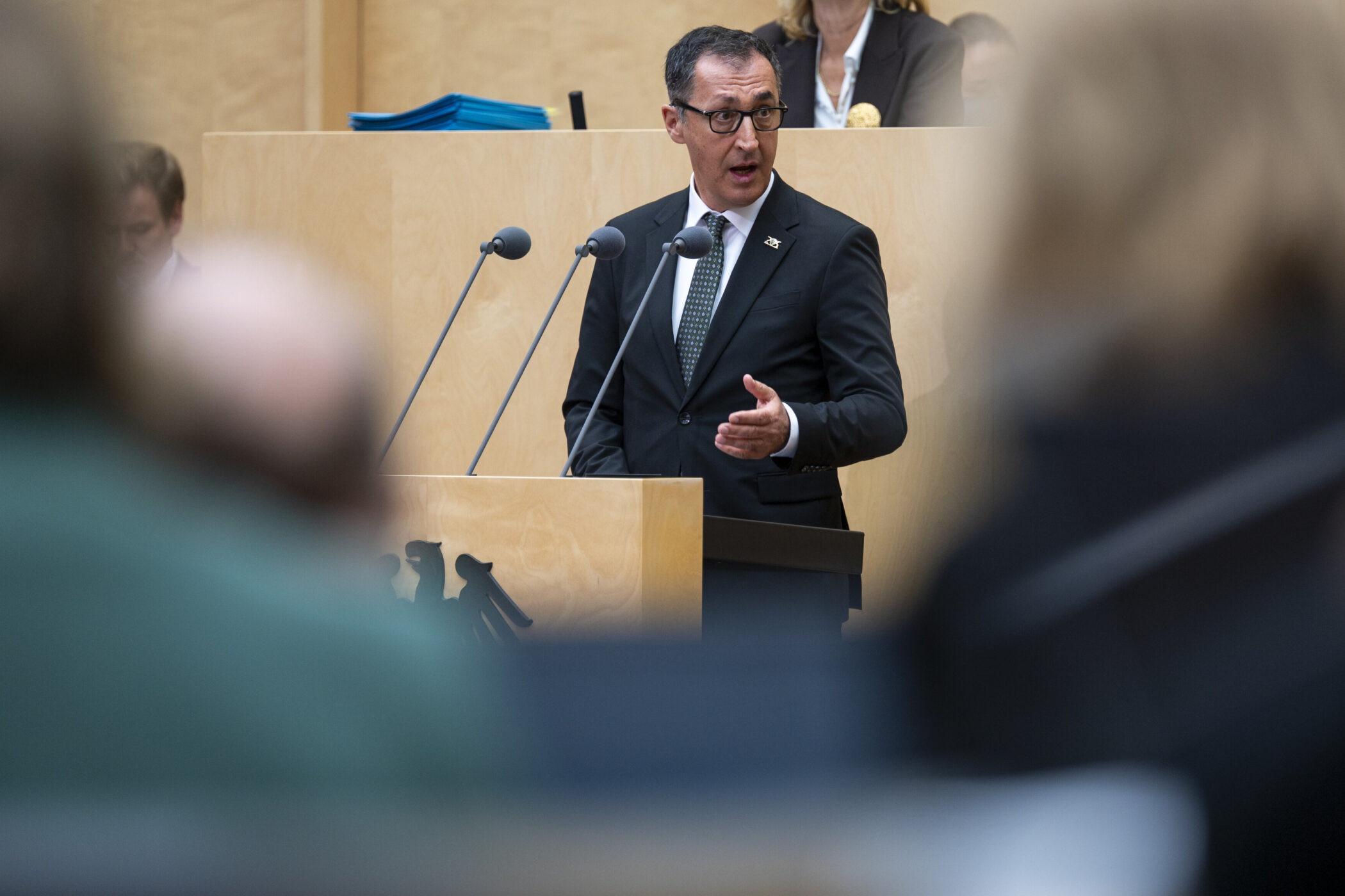 Mann im Anzug spricht am Rednerpult mit Mikrofonen, unscharfe Personen im Vordergrund.