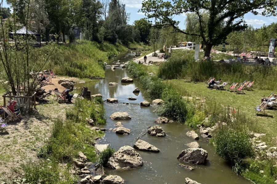 Kleiner Fluss mit großen Steinen, umgeben von Gras, Menschen auf Liegestühlen.