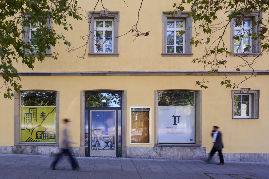 Gelbes Gebäude mit fünf Fenstern; zwei Personen gehen vorbei.