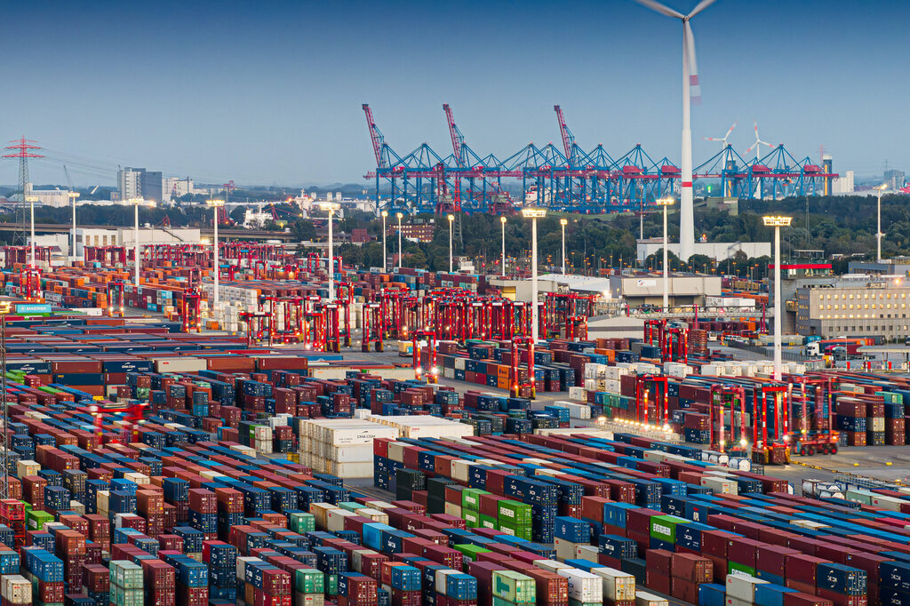 Containerstapel und Kräne im Hafenbereich, im Hintergrund Gebäude und Windkraftanlage.