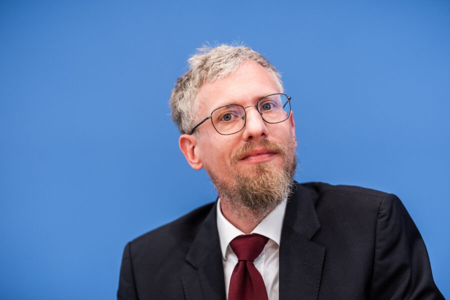 Mann mit Brille und Anzug vor blauem Hintergrund.