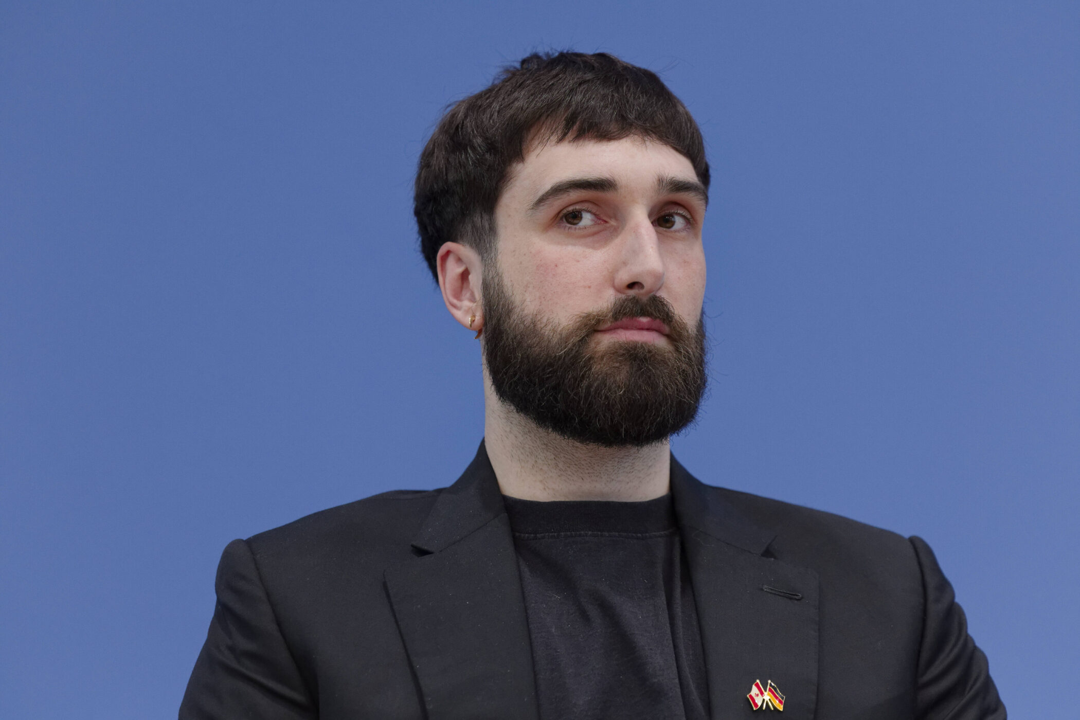 Ein Mann mit Bart vor blauem Hintergrund, trägt schwarzen Blazer.