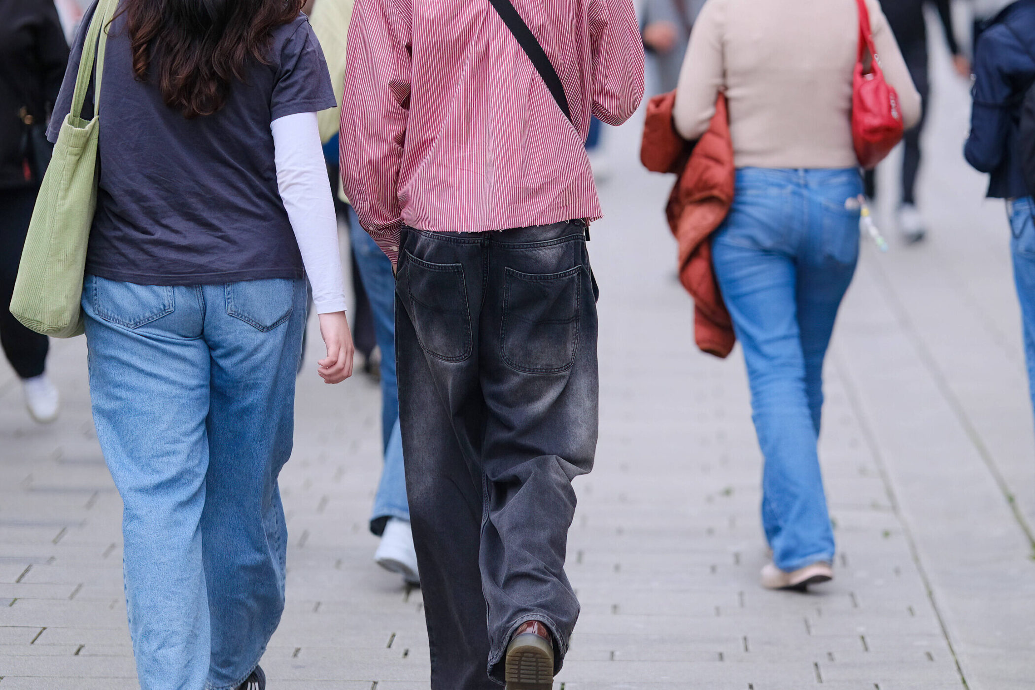 Drei Personen von hinten, gehen auf Gehweg, tragen Freizeitkleidung und Taschen.