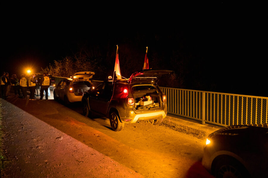 Zwei Autos mit offenen Kofferräumen auf Brücke bei Nacht, mehrere Personen daneben.