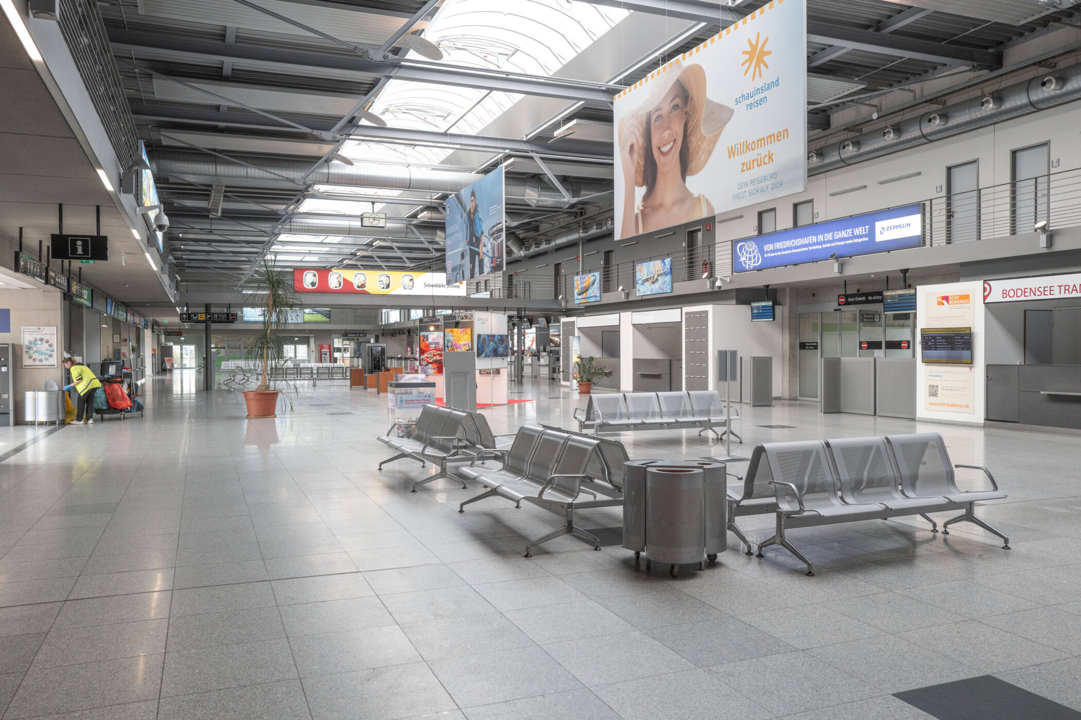 Leerer Flughafen-Terminal mit Sitzbänken und Werbetafeln, eine Person rechts.