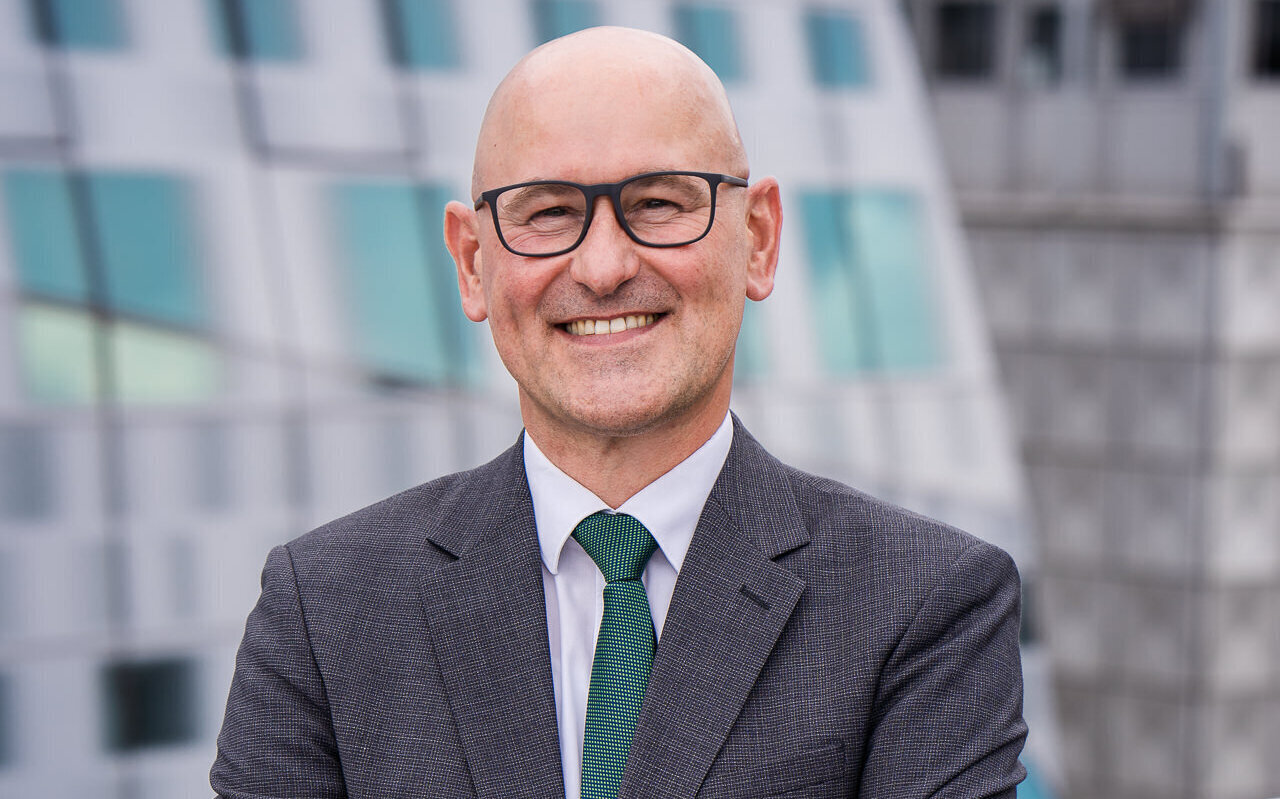 Mann mit Brille und Anzug lächelt, Hintergrund aus Glas und Metall.