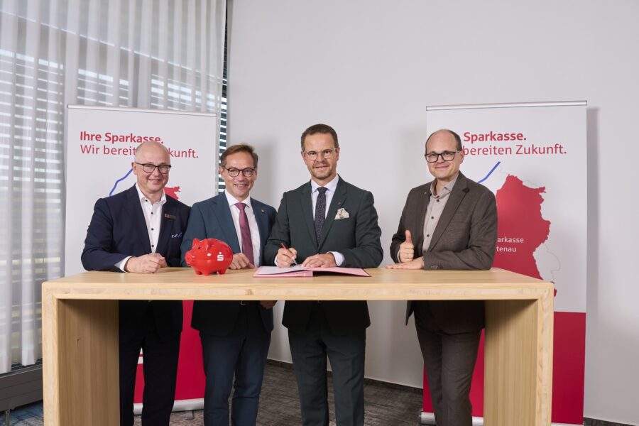 Vier Männer stehen an einem Tisch, einer unterschreibt ein Dokument. Zwei Banner im Hintergrund.