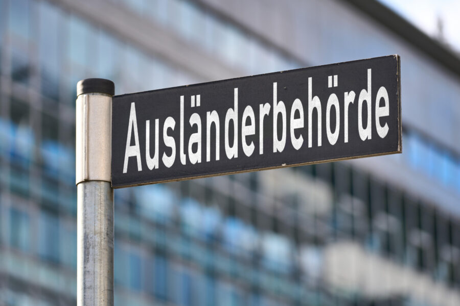 Schild mit der Aufschrift „Ausländerbehörde“ vor unscharfem Gebäudehintergrund.