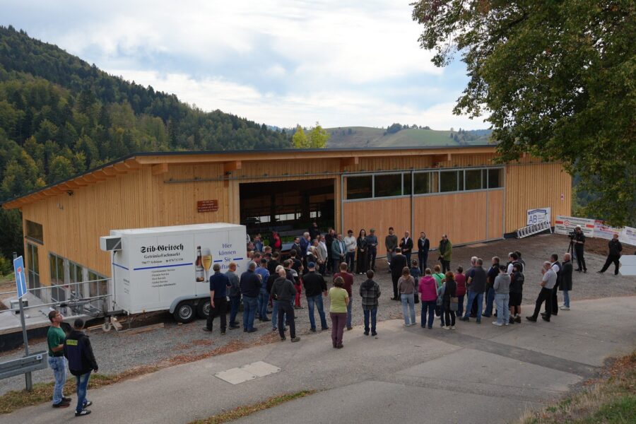 Personengruppe steht vor einem Holzgebäude mit Anhänger. Wald im Hintergrund.