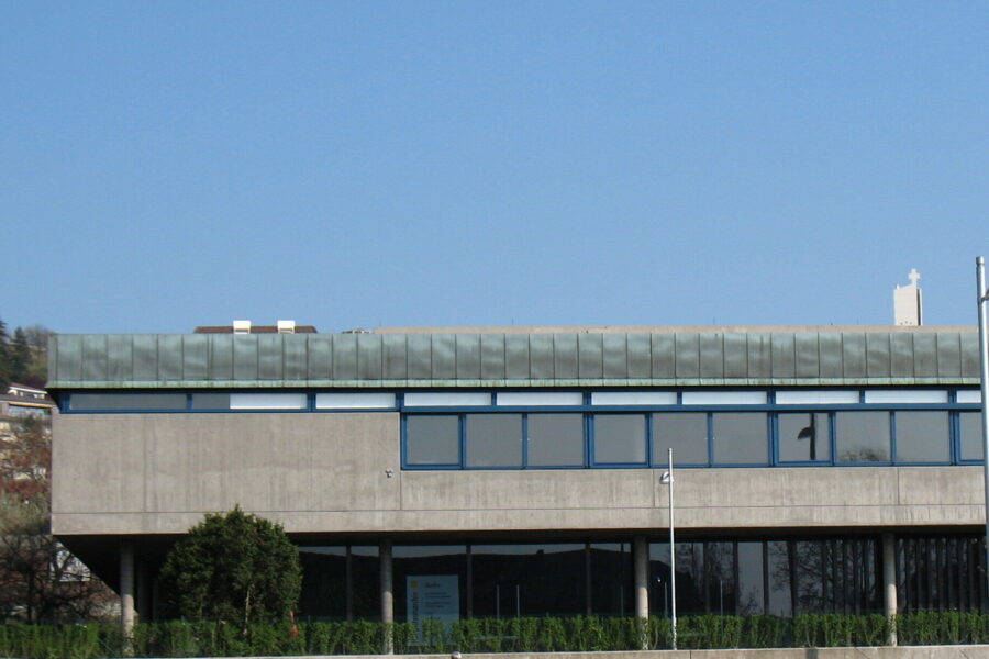 Modernes Gebäude mit großen Fenstern, flacher Dachkante, im Vordergrund Sträucher.