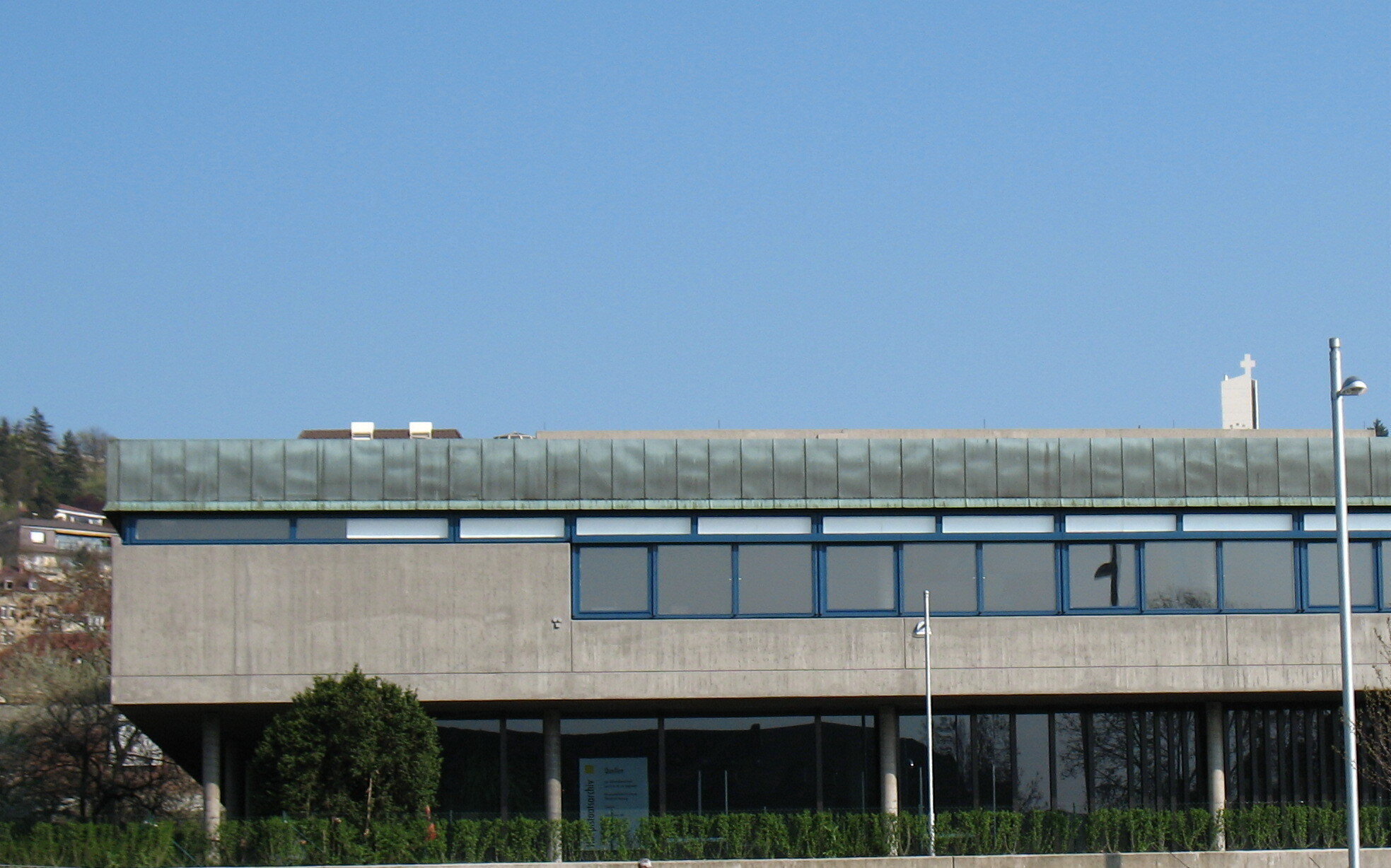 Modernes Gebäude mit großen Fenstern, flacher Dachkante, im Vordergrund Sträucher.