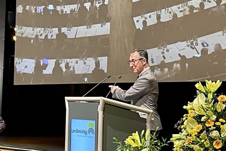 Ein Mann steht am Rednerpult vor einem großen Bildschirm, rechts Blumenarrangement.