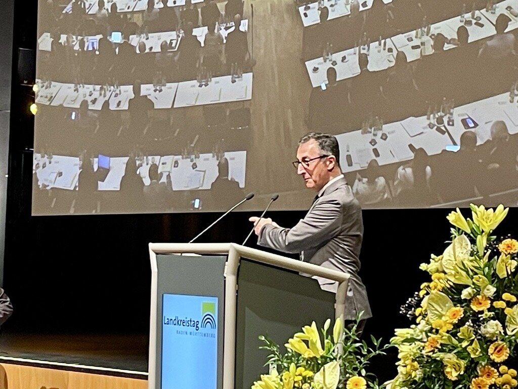 Ein Mann steht am Rednerpult vor einem großen Bildschirm, rechts Blumenarrangement.