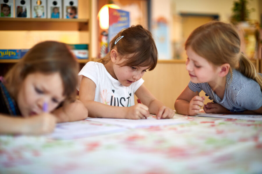 Drei Kinder malen an einem Tisch in einem hellen Raum.
